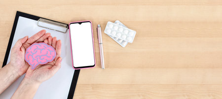 Female hands holding brain figure, smart phone, clipboard and pills on wooden desk.の写真素材