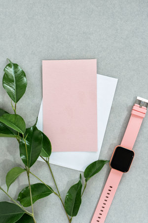 Top view of empty pink card with white envelope, watch and leaves, on gray background.の写真素材
