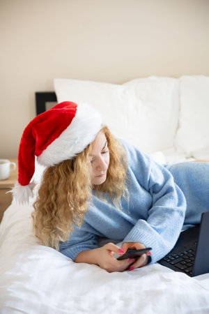 Woman in santa claus red cap with smartphone and laptop in  bed.