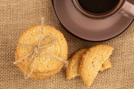 Stack of oatmeal cookies with sesame seeds tied with string on burlap cloth on wooden table. Cup of coffee. Top horizontal view.の写真素材