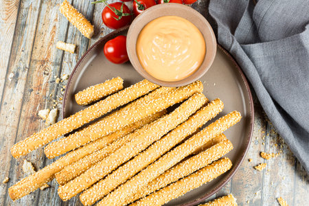 The bread sticks with sesame seeds on brown ceramic plate, cheese sauce, cherry tomatoes and gray...
