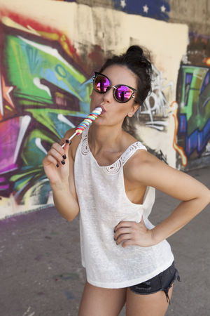 Cute woman with sunglasses eating sexy lollipop and posing in front of graffiti wall.の写真素材