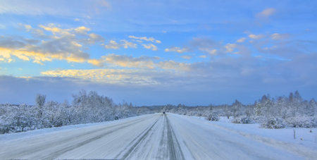 Winter winding  snowy highway leading through a mountain valley landscape, with forest covered, blurred from hight speed drive. Evening twilight, golden sunset clouds - concept of safety in winter drivingの写真素材