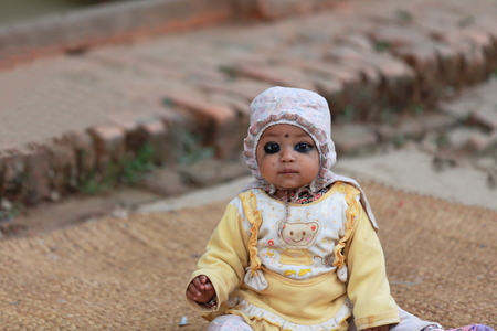 GODAWARI, NEPAL - OCTOBER 15: Eye painted little baby sits on a mat at the entrance to an old newar style house in a street of Godawari. Lalitpur distr.-Bagmati zone-Nepal.のeditorial素材