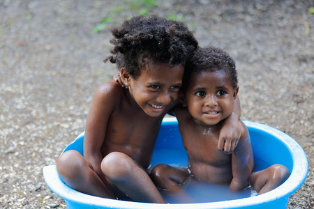 Lamen Bay, Epi island, Vanuatu-October 4, 2014: Little girl and boy of the Ni-Vanuatu people take a bath in a big plastic washbasin placed in front of the door of their home in the beginning of sunsetのeditorial素材