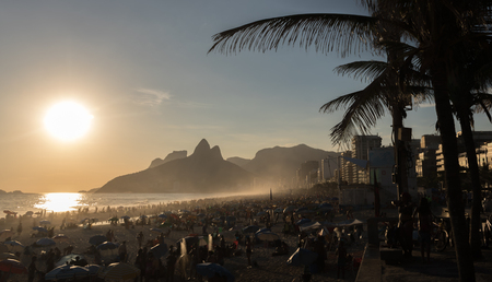 The sun is decending on the beach in Rio de Janeiro. The beach is crowded with people waiting for a stunning sunset. There are silhoettes of palm trees in the foreground and mountains in the background.のeditorial素材