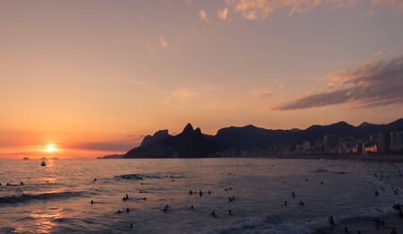 The sun is decending down below the horizon which is giving off a beautiful orange, red, and yellow light. There are sillhoettes of a crowd of people on the beach and the Two Brother Mountain in the background.のeditorial素材