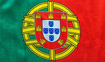 A Portuguese flag is pictured as it lays on a flat surface. The Coat of Arms in between the bold red...