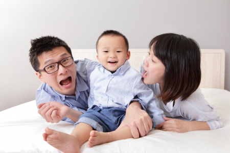 A happy family with funny facial expression lying on white bed in the bedroom with gray wall for copy space, asian peopleの写真素材