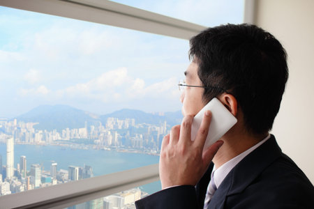 Young business man talking on smart phone in office and look city through window, asia, hong kong,...