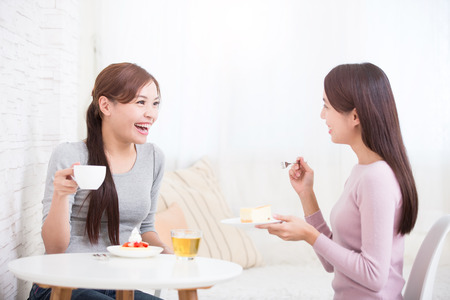 Two happy young female friends with coffee cups and cakes enjoying a conversation in the living room at home, healthy lifestyle concept, asian beauty