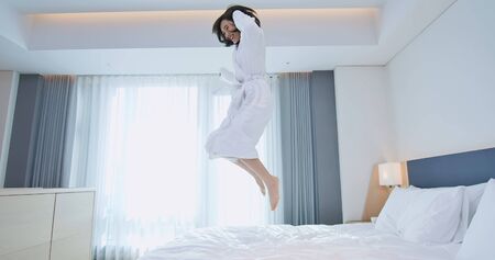 Young beautiful female having fun and jumping with bathrobe on bed in hotel roomの写真素材