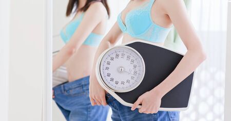 Girl lose weight successfully and show her big jean before in front of mirror with scaleの写真素材