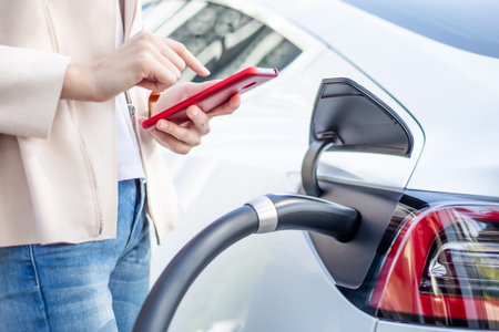 Close up of woman use smart phone while waiting and power supply connect to electric vehicles for...