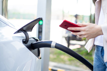 Close up of woman use smart phone while waiting and power supply connect to electric vehicles for...