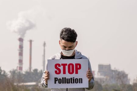 asian man hold stop pollution sign and wears protective n95 mask against air pollution standing in front of factoryの写真素材