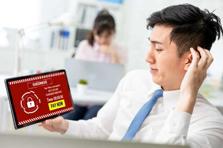 asian worried young businessman looking at tablet pc with ransomware attack words on the screen in officeの写真素材