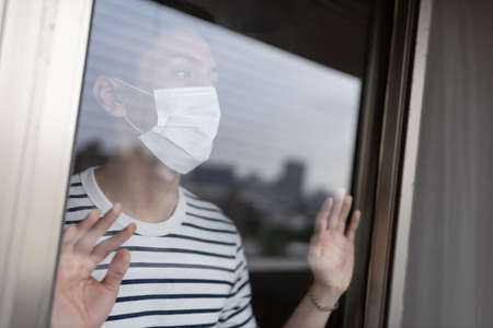 asian young man with face mask stay isolation at home for self quarantine due to an epidemic of COVID-19 and he looks out by windowsの写真素材