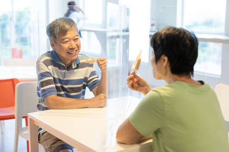 Asian elder couple man and woman sitting in restaurant eating food with table shield to protect infection from coronavirus covid-19の写真素材