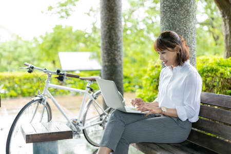 Asian businesswoman wear face mask and use laptop to work with bicycle in the parkの写真素材