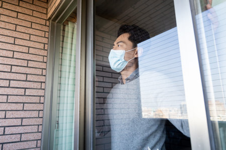 close up of asian young man wearing face mask stay isolation at home for self quarantine due to epidemic of COVID19-he looks out the windowの写真素材