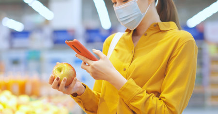 close up of asian young businesswoman wear face mask who is shopping for fresh organic fruits and scanning barcode with smart phone at supermarket during virus epidemic outbreakの写真素材