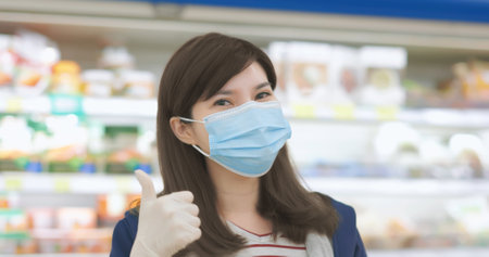 close up of smiling asian homemaker is wearing face mask with gloves then shows thumbs up gesture at grocery store during virus epidemic outbreak-look to youの写真素材