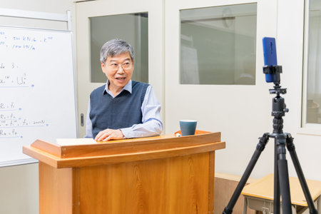 Asian mature male professor teach Engineering online through mobile phone in classroom at graduate...
