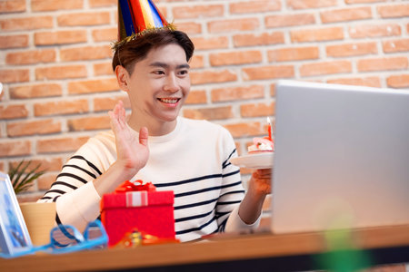 asian young man wearing birthday hat has video chat online by laptop while celebrating and giving cake for his friendの写真素材