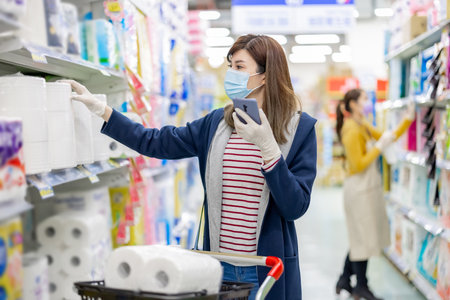 asian homemaker wearing face mask and gloves is using smart phone to check shopping list while buying paper towel at grocery store during virus epidemic outbreakの写真素材