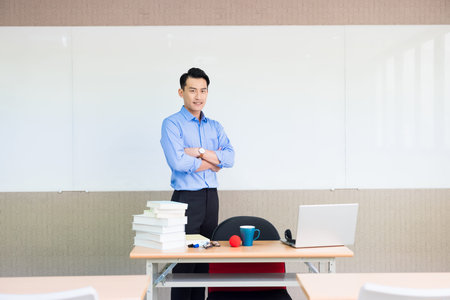 asian young male teacher smiles at you with cross arm standing front of whiteboard in classroomの写真素材