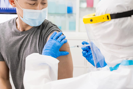 close up of nurse wearing gloves and isolation gown is making a vaccination in the shoulder of asian...
