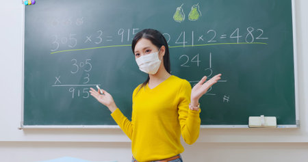 asian elementary school female teacher wearing face mask teach math online through webcam in...