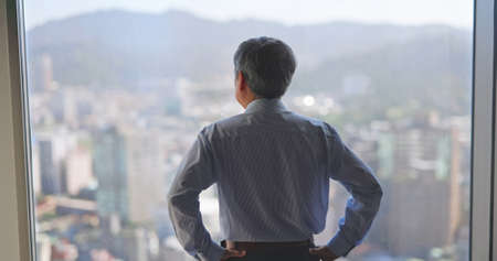 asian senior businessman is looking at city view through windowの写真素材