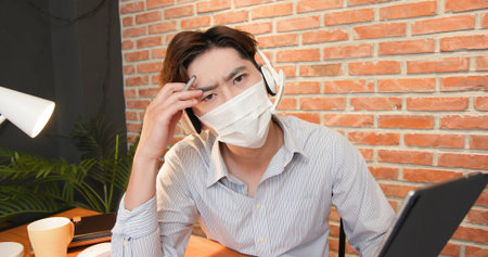 telework concept-Asian man wearing face mask and headset is very upset to get some trouble about his work on a video meeting at homeの写真素材