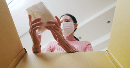 asian young woman wearing surgical face mask is opening online shopping parcel cardboard box during the epidemic prevention periodの写真素材