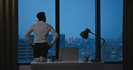 worried asian young businessman is looking out of windows while working overtime in office at nightの写真素材
