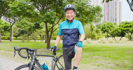 Smiling asian old man wearing helmet and touch his knee with a bicycle in the parkの写真素材