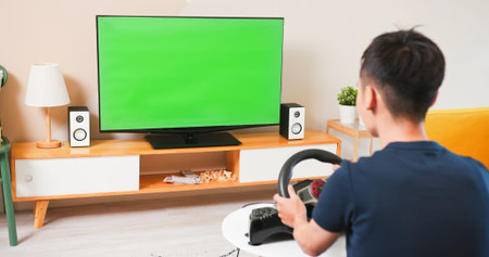 Asian gamer boy is sitting on couch playing racing game with green chroma key TV screen at home-he...