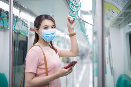 Asian business woman use a smartphone with surgical mask face protection while commuting and smile at you in the metro or trainの写真素材