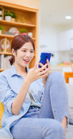 young Asian woman using smartphone checking social media feeling happy smiling while sitting on sofa...