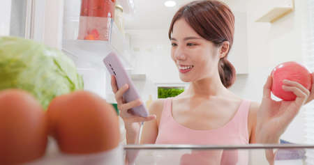asian woman use smartphone making list of food standing with opened fridgeの写真素材