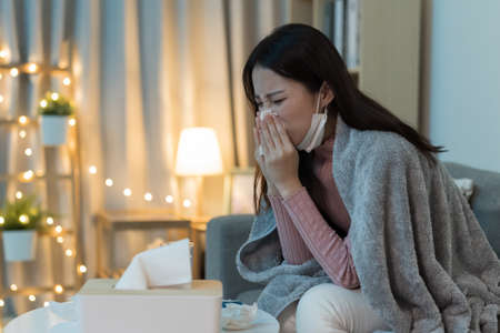 Sick asian woman is blowing running nose and sneezing in tissue at homeの写真素材