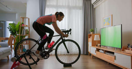 side view of asian woman watching green screen television while riding exercise bike in living room at homeの写真素材