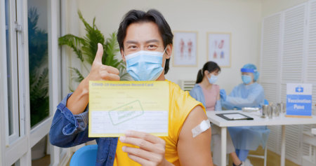 asian man wears face mask showing COVID-19 vaccination record card with thumb up gesture in hospital