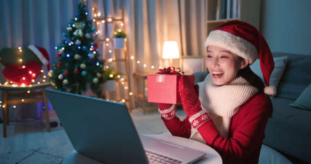 asian young woman wearing christmas clothes has video chat on laptop and shows present box to her friendの写真素材
