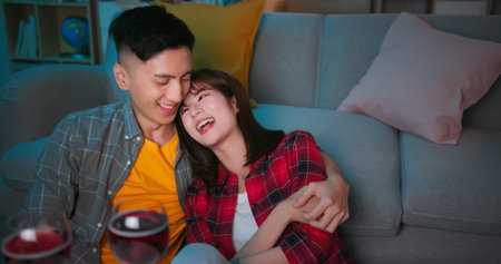 asian couple sitting on floor smile with red wine while celebrating anniversary at night in living...