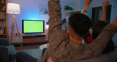 back view of Asian young excited couple are watching sport games on TV with green chroma key screen and having fun at night in the living room-raising armsの写真素材