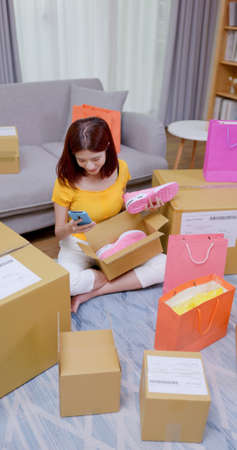 high angle view of happy asian woman receive packing parcel after online shopping at home-she check order list on mobile phone with opening delivery boxes or bagsの写真素材