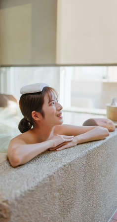 smiling asian young woman lying on the bathtub is relaxing in hot spring with towel on headの写真素材